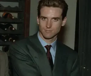 A man in a formal green suit and red tie, standing indoors near a wine rack, looking at the camera with a slight smile.