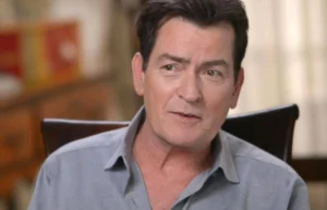 Charlie Sheen is sitting in a chair, laughing while talking.