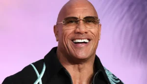 Bald man with glasses smiling brightly in a black embroidered jacket against a soft purple background.