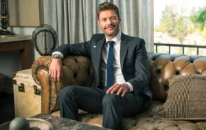 Ryan Seacrest is sitting on the couch, smiling.