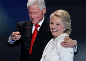 Bill Clinton is looking at something with his hand on the woman's shoulder and gesturing.