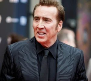 A man in a stylish black patterned suit and tie smiling at an event, with a blurred background.