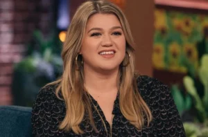Kelly Clarkson is smiling while sitting on the couch.