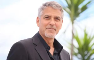 George Clooney is smiling while standing outside in front of the plants.