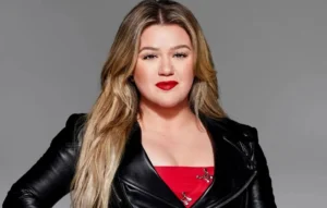 Kelly Clarkson is smiling while wearing a black coat.