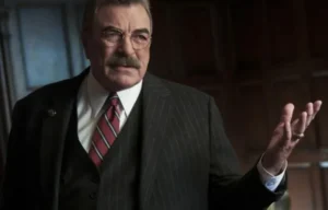 Tom Selleck is wearing a black coat and gesturing with his hand while talking.