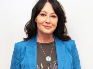 Shannen Doherty is laughing, wearing a blue coat and a chain around her neck.
