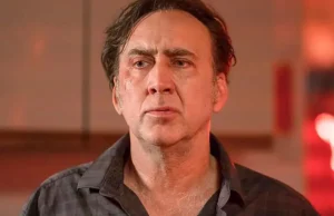 A middle-aged man with a serious expression, wearing a dark checkered shirt, standing indoors with a warm light in the background.