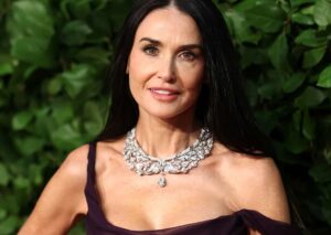 Woman in a dark purple off-shoulder dress wearing a large diamond necklace, standing in front of green leafy background.