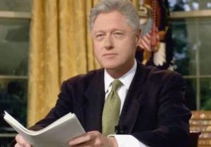 Bill Clinton is sitting in the room with a paper in his hand.