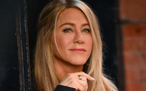 Close-up of a woman with long blonde hair, light blue eyes, and a calm expression, wearing a black outfit and a gold ring, looking slightly upward.