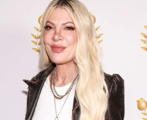 Tori Spelling is smiling while wearing a black coat.