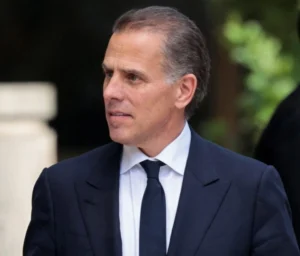 Hunter Biden is looking to the side, wearing a black coat and black tie.