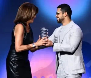 Woman in a black dress receiving an award from a man in a light gray suit on stage.