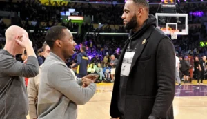 Two men talking courtside during a basketball game, with players warming up in the background