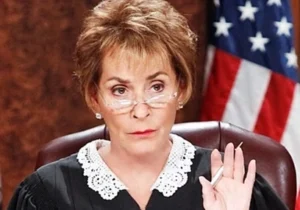 Judge Judy is sitting in a chair, holding a pen in her hand, gesturing for silence.