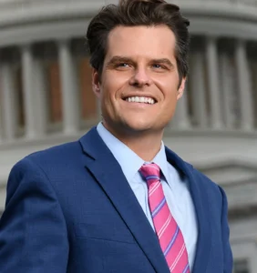 Matt Gaetz is laughing while wearing a blue coat and a white building is visible in the background.