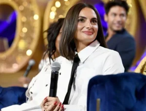 A woman wearing a white shirt and black tie smiles while holding a microphone on stage, with people and a sparkling background behind her.