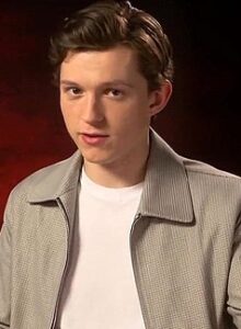 Man wearing a light gray jacket and white t-shirt speaking during an interview with a red background.