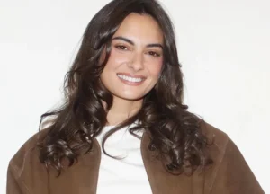 A smiling woman with long wavy dark hair wearing a white top and brown jacket poses against a plain background.