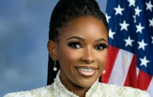 Jasmine Crockett is laughing while wearing white clothes and the American flag stands behind her.