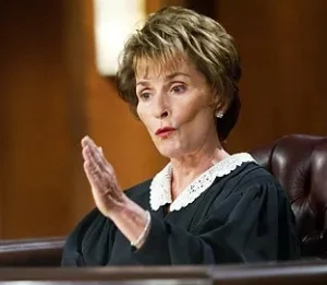 Judge Judy is sitting in a chair, wearing a black coat, talking.