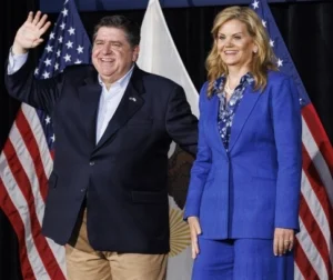 JB Pritzker stands with his wife, laughing, and the American flag behind him.