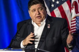 JB Pritzker is speaking into a microphone and an American flag stands behind.