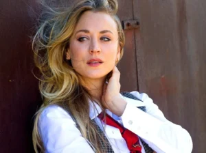 A woman with long blonde hair wearing a white shirt, red tie, and gray vest, posing outdoors with her hand on her neck.