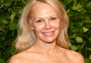 Pamela Anderson is smiling with her hair open and green plants standing in the background.