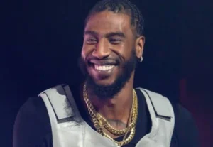 Iman Shumpert is laughing, wearing a shirt and a chain around his neck.