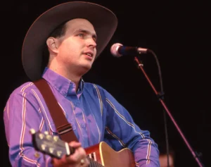 Garth Brooks stands in front of the mic, holding a guitar.