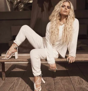 Blonde woman in a stylish cream colored loungewear set with long wavy hair, sitting on a wooden bench and wearing white strappy high heeled sandals.