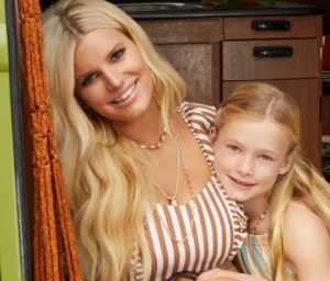 Smiling blonde woman in a striped outfit posing with her young daughter, both wearing casual jewelry and looking happy.