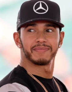 Lewis Hamilton is seen looking at a sideline wearing a black shirt and black hat.