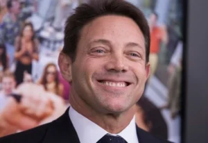 Jordan Belfort is laughing in a black coat with pictures of girls on the back.
