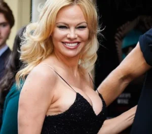 A smiling blonde woman in a black dress with thin straps, looking towards the camera while holding someone’s arm.