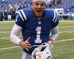 Pat McAfee stands on the ground, wearing his uniform and holding his helmet.